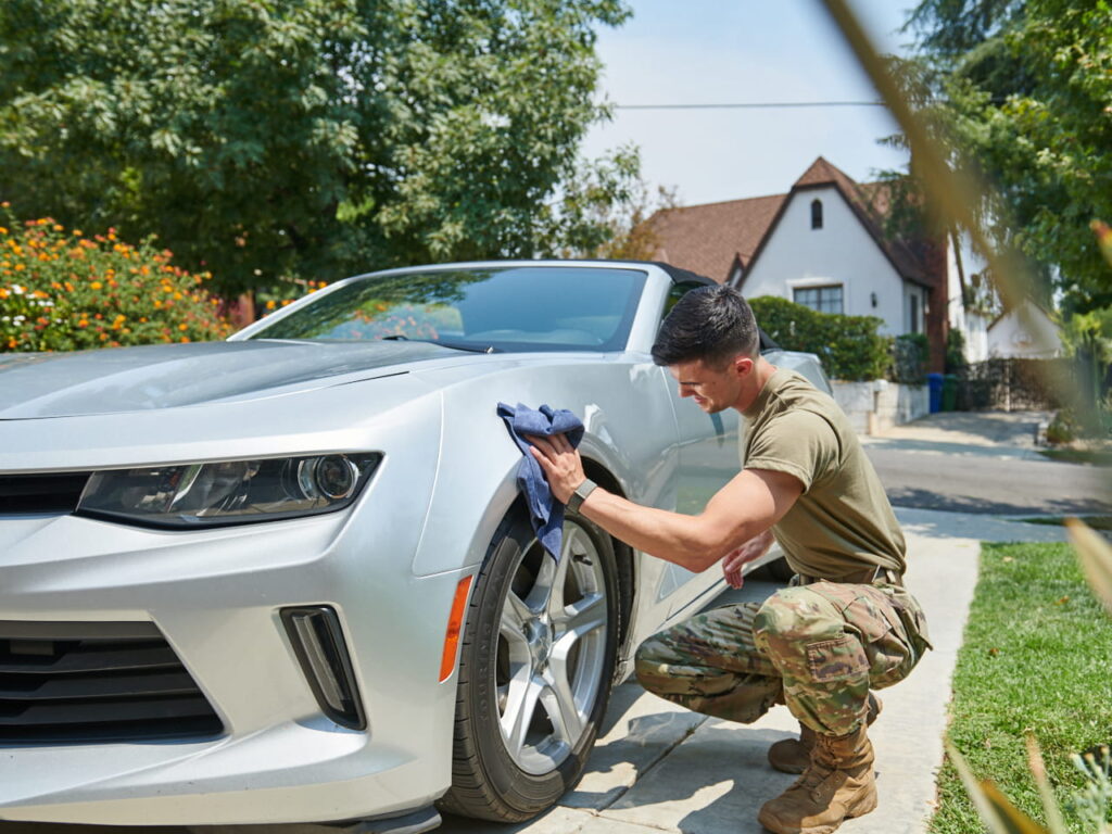 Military man in camo pants and olive t-shirt wiping the fender of a sports car with a rag.